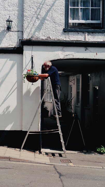 A man on an aluminium stepladder tending to a hanging flower basket outside a whitewashed building in Lyme Regis.