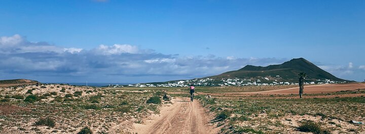A cyclist rides a dirt track through dry fields toward the village of Soo and nearby volcanic hills beneath a wide blue sky.