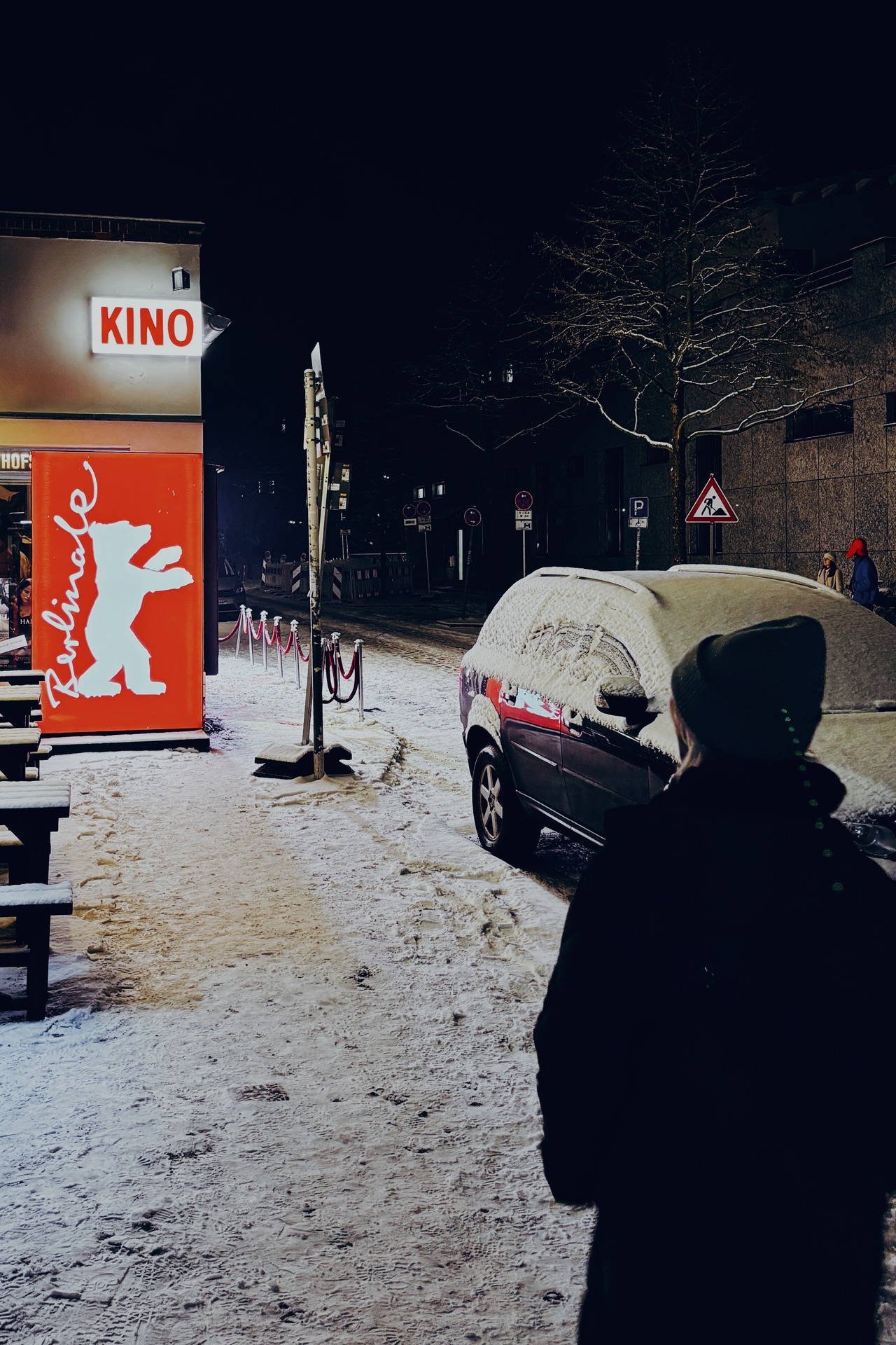 Snowy night street scene in Berlin beside a red Berlinale sign, with a snow-covered parked car and a person in the foreground.