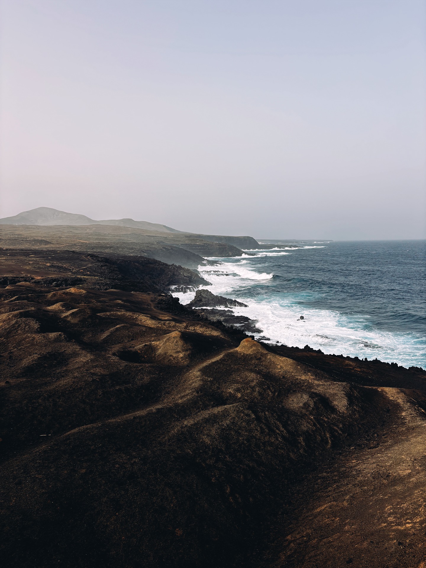 Dark volcanic coastline under a hazy sky, with white surf rolling into rocky coves along the shore.