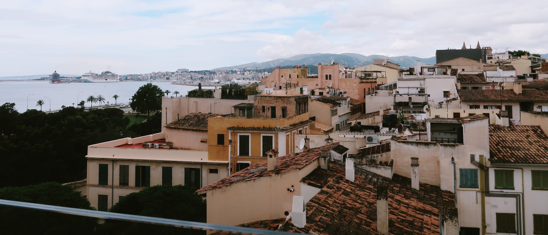 Panoramic view over the terracotta rooftops of Palma's old town, with the harbour and sea visible to the left and mountains along the horizon.